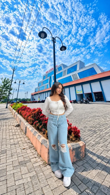 Woman in a white crop top, ripped light blue jeans and white sneakers standing on a paved street under a cloudy blue sky