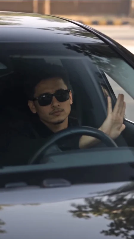 Man wearing sunglasses sitting in the driver's seat of a black car and waving through the windshield
