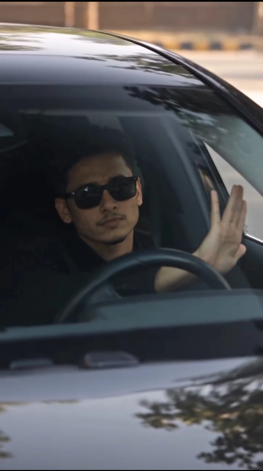 Man wearing sunglasses sitting in the driver's seat of a black car and waving through the windshield